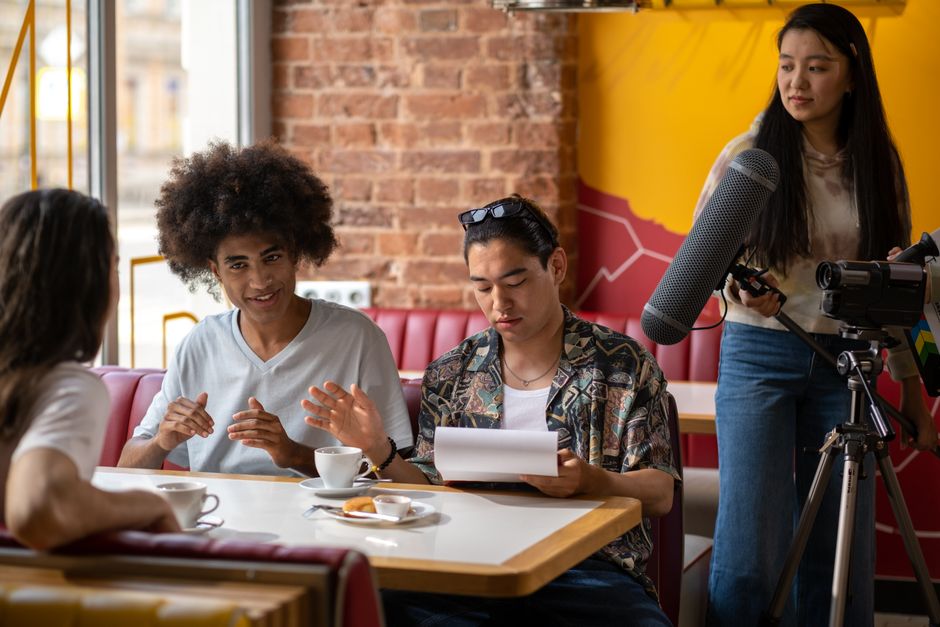 A diverse group of filmmakers working indoors in a cafe setting, capturing a scene with a camera and microphone.