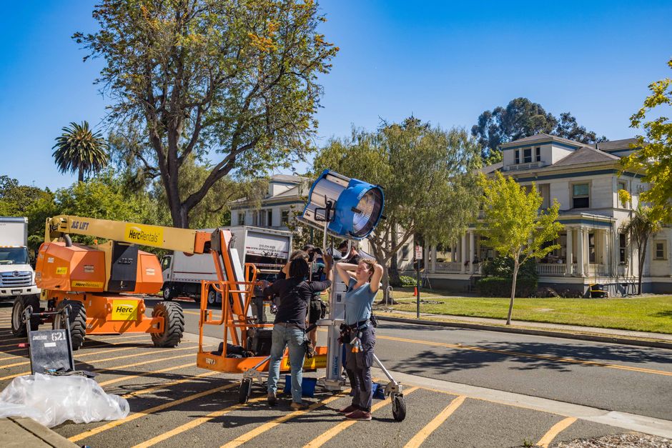 Film crew setting up lighting equipment outdoors with lift and tree backdrop.