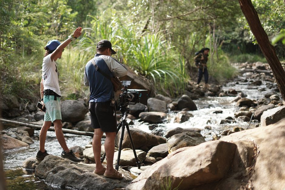 Film crew operates camera equipment by a serene forest stream, capturing nature's beauty.