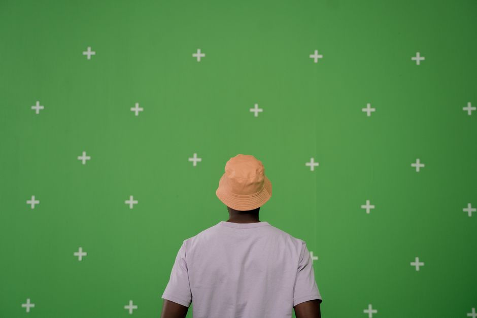 A person in a bucket hat stands before a green screen backdrop with crosses.