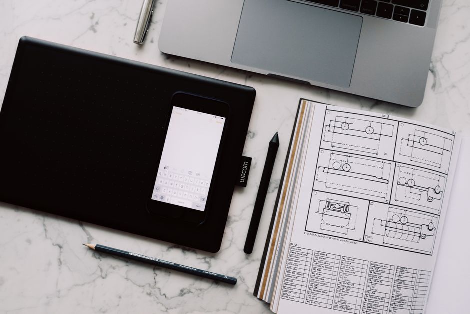 A modern workspace featuring a drawing tablet, smartphone, laptop, and design documents on a marble desk.