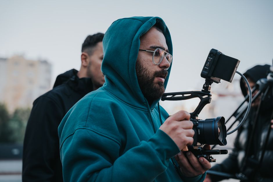 Filmmaker in blue hoodie operating a camera outdoors with a screen monitor attached.