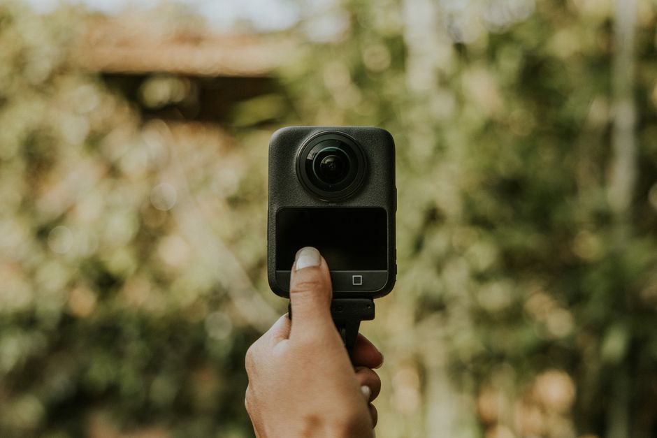 Person holding a 360 camera outdoors with blurred green background.