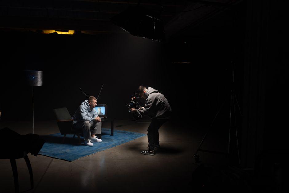 Film production shoot indoors with cinematographer filming an actor sitting, studio lighting.