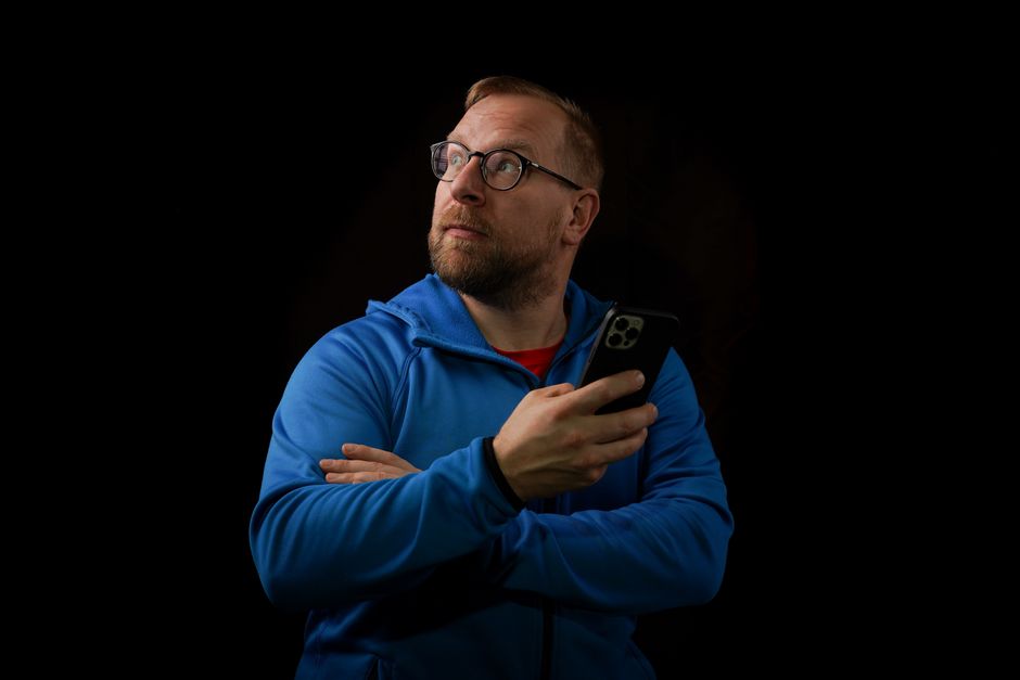 A thoughtful man in a blue jacket holds a smartphone against a black background, looking upwards.