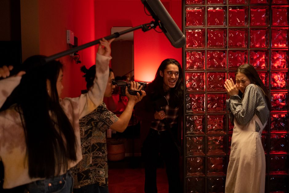 A group of teenagers filming a playful horror scene indoors with a boom mic and camera.
