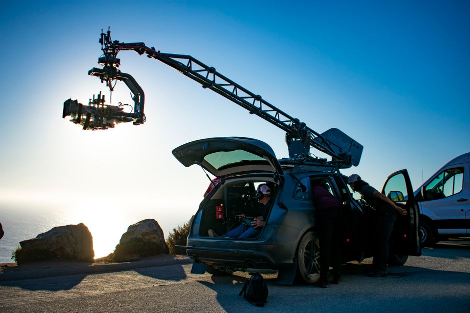 A film crew operates a camera crane mounted on a vehicle at a cliffside location during sunset.