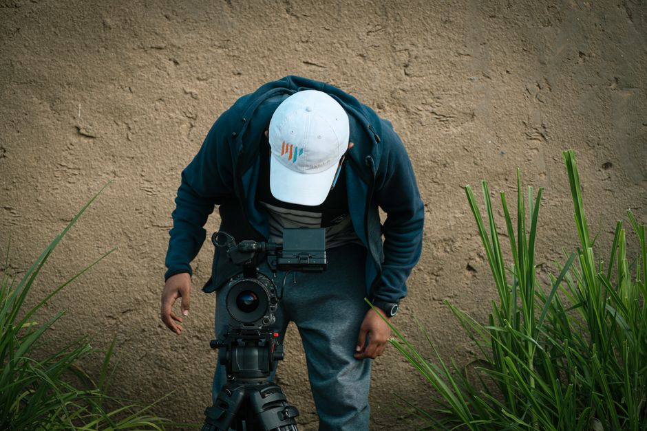 A man adjusts a camera setup outdoors near a grassy wall.