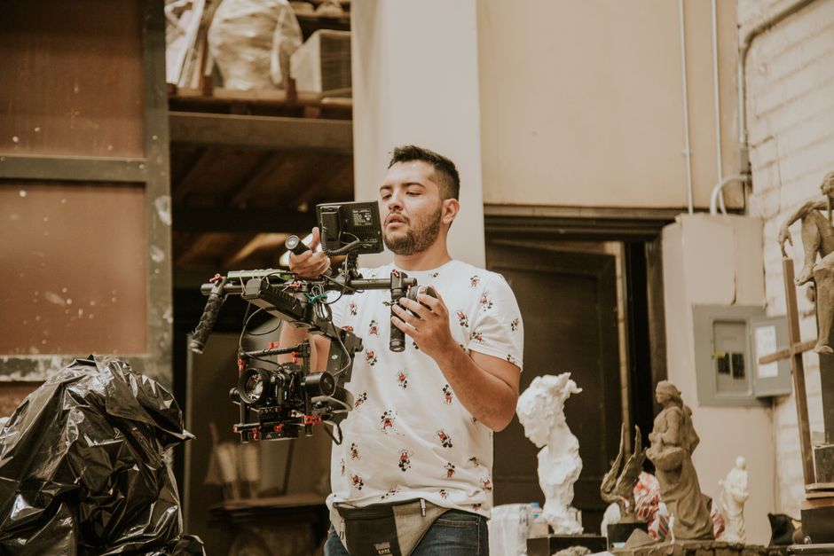Male videographer holding a professional camera in an art studio surrounded by sculptures.