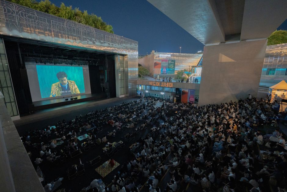 A large audience gathers outdoors at night to watch a movie screening under a bridge.