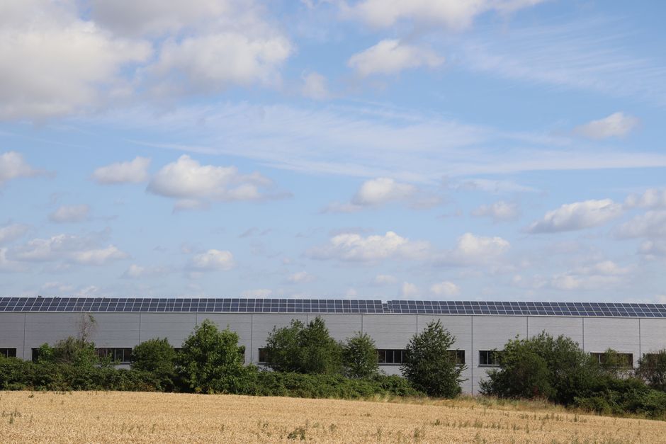 A countryside warehouse with solar panels and fields under a blue sky with clouds.