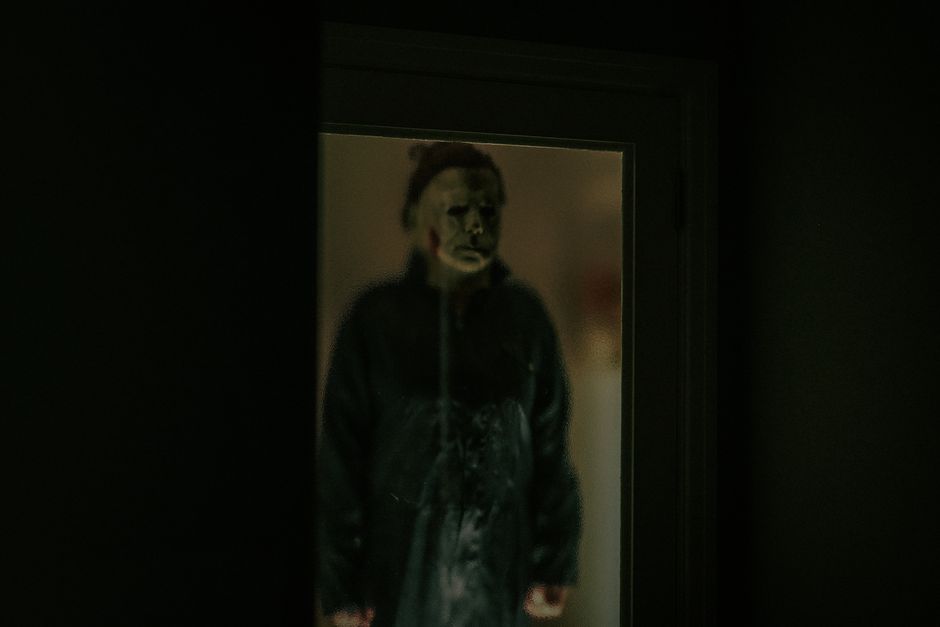 A chilling figure in a Halloween mask is reflected in a dark window, creating an eerie atmosphere.