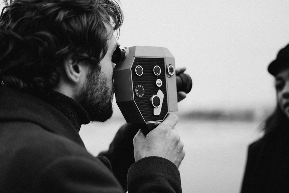A black and white photo of a man with a retro video camera filming a woman outdoors.