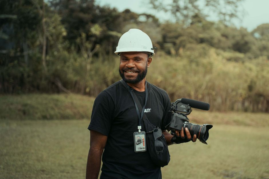 A cheerful cameraman in a hard hat holding a video camera in a scenic outdoor setting.