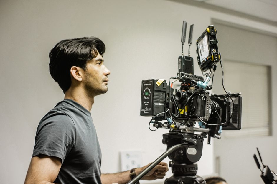 A professional videographer in a gray shirt operates a high-end camera setup indoors, capturing footage in a studio environment.