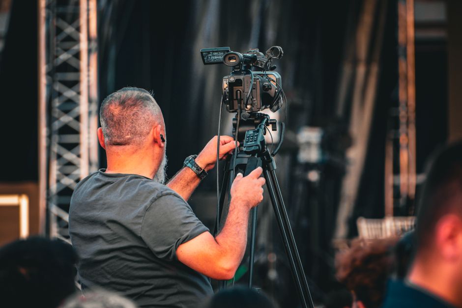 A professional cameraman operating a video camera on a tripod during an outdoor event, captured from behind.