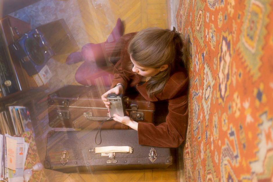 From above of faceless female in retro outfit sitting on floor near vintage suitcases and considering film camera
