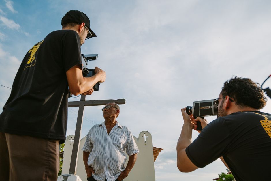 Two filmmakers document an elderly man in an outdoor setting, capturing the moment with professional cameras.