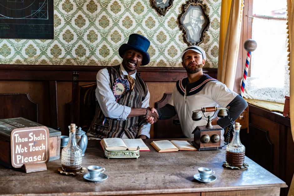 Two men in vintage costumes shake hands in a themed historical setting.
