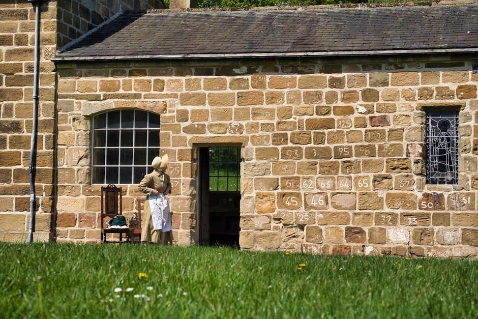 Outdoor scene of a historic stone house facade with mannequin in period attire; rural setting.