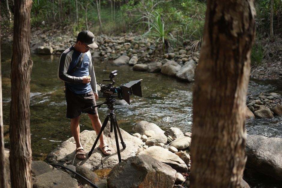 A cameraman operates equipment beside a stream in a forest setting in Mati, Philippines.