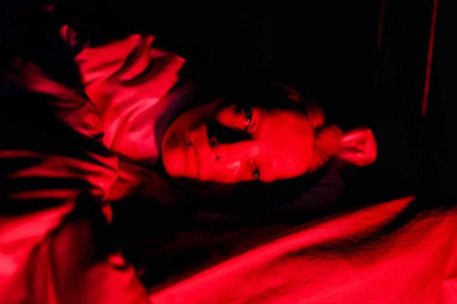 A person in bed is illuminated by bold, red lighting creating a vivid, dramatic portrait.