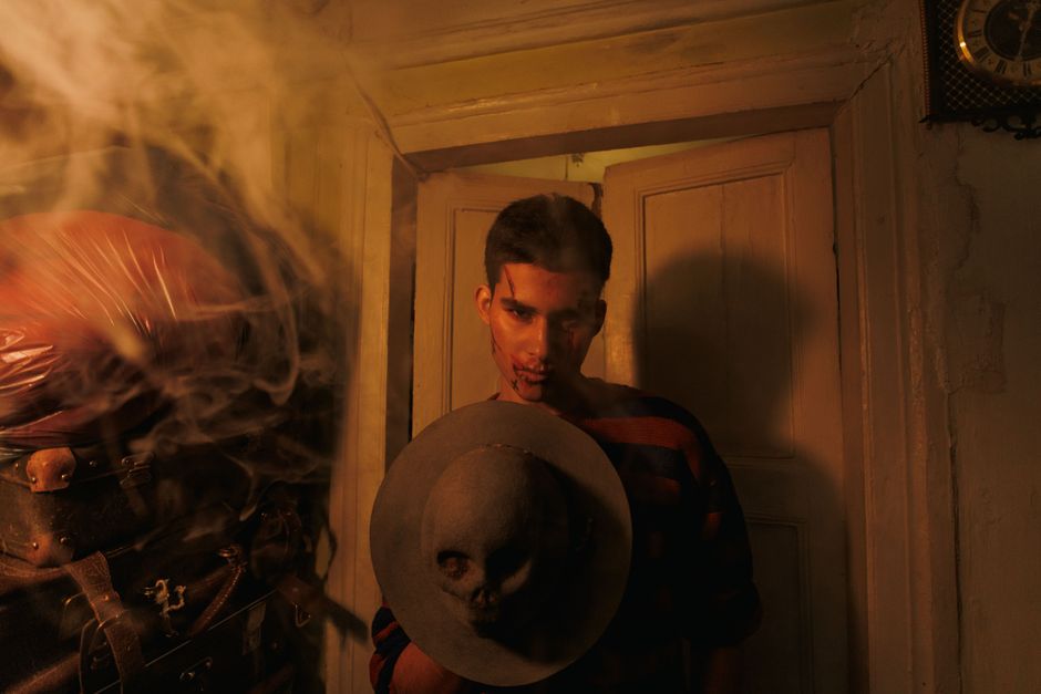 A man in a dimly lit room holds a hat adorned with a skull, surrounded by smoke.