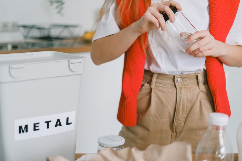 Crop anonymous female standing at table with different litter and bucket for metal trash and unscrewing glass jar