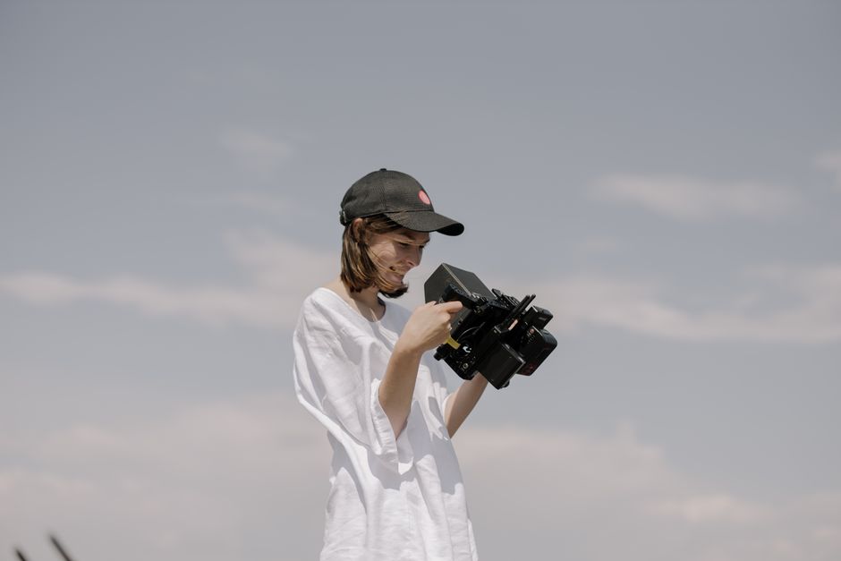 A woman smiling while holding and examining a professional video camera outdoors.