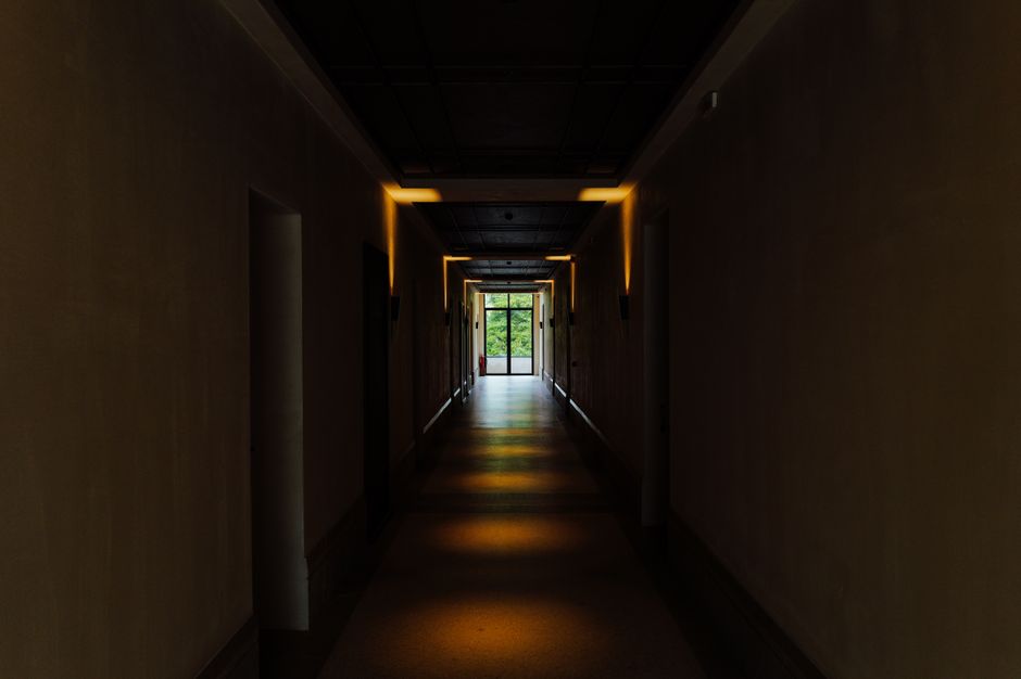 A shadowy indoor hallway with lights leading to a bright exit, creating a sense of mystery.