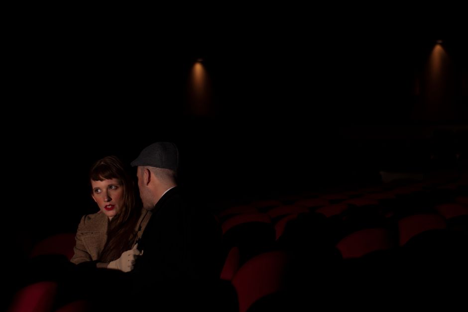 A couple sitting closely in a dimly lit movie theater, whispering to each other.