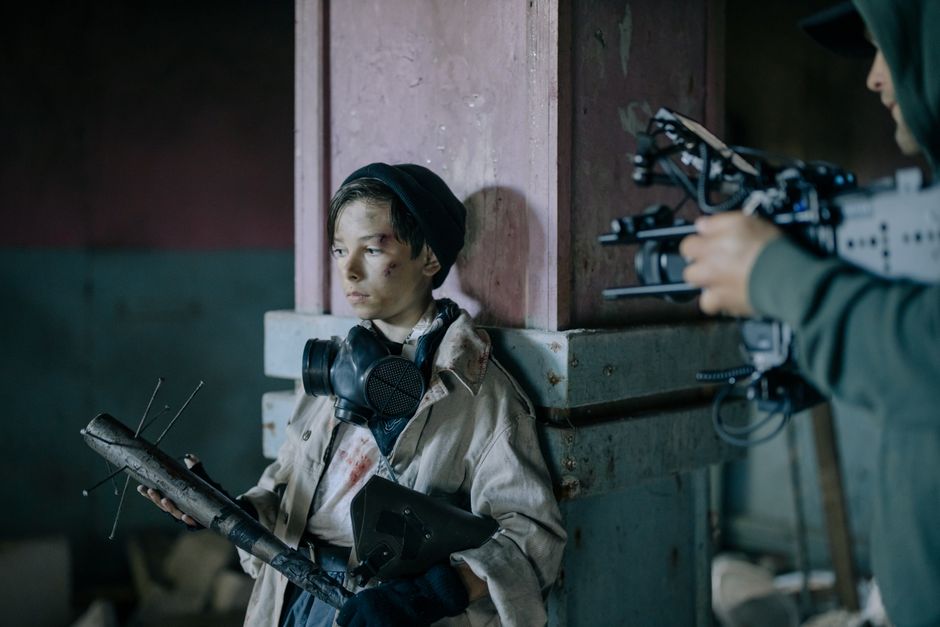 A young boy is photographed in a post-apocalyptic setting, holding a nail bat and wearing a gas mask.