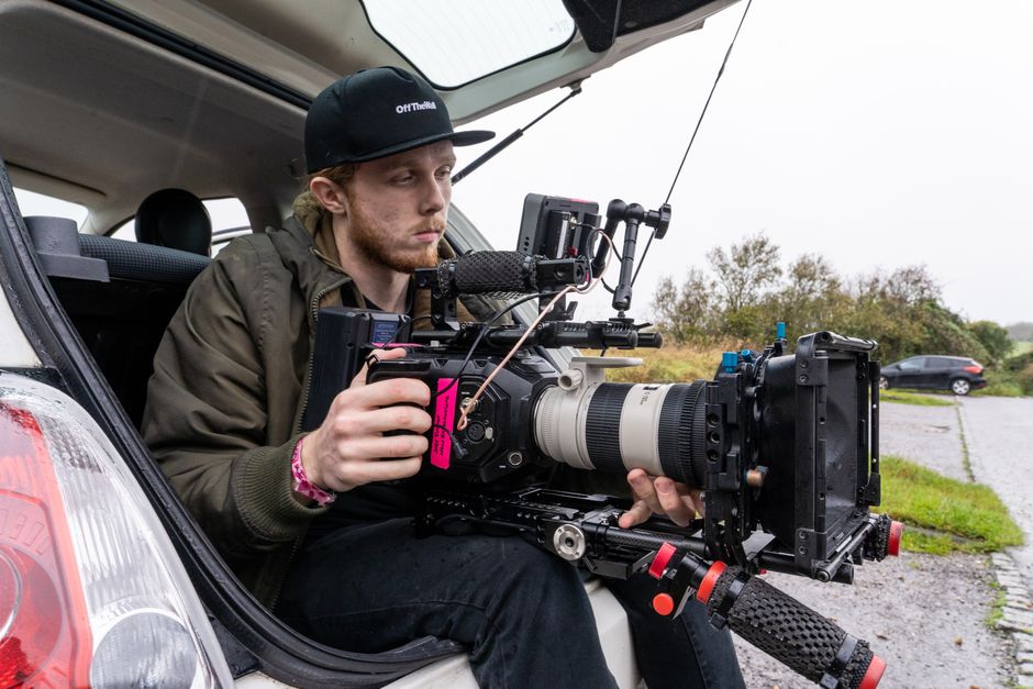 Cameraman seated with large video camera, filming from car trunk in daylight outdoors.