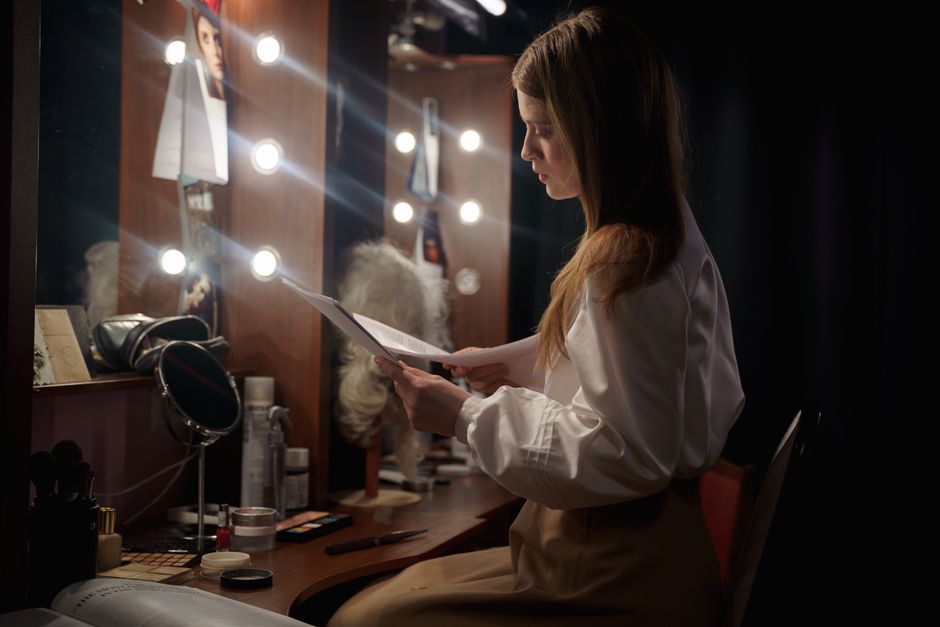 A woman in a dressing room reading a script, surrounded by makeup and lights.