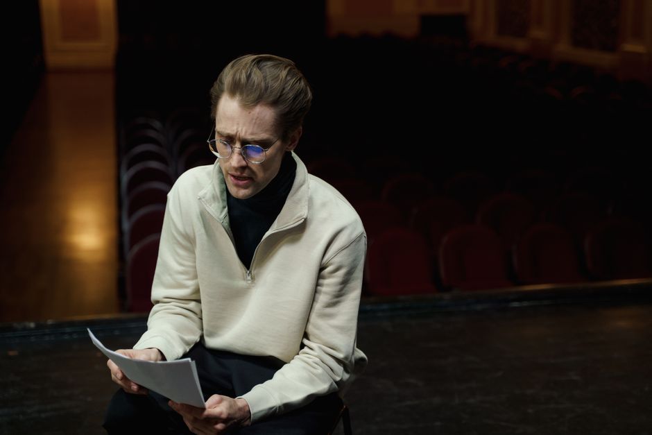 An actor rehearses lines on a theater stage, conveying focus and dedication in a dimly lit setting.