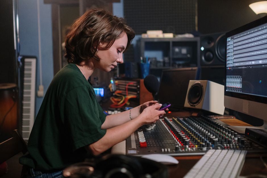 A woman in a green t-shirt uses her smartphone while sitting at a sound mixer in a recording studio.