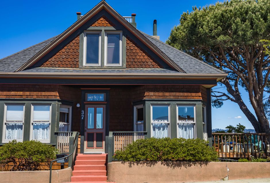 A picturesque vintage house with red stairs, green trims, and bay windows set against a clear blue sky.