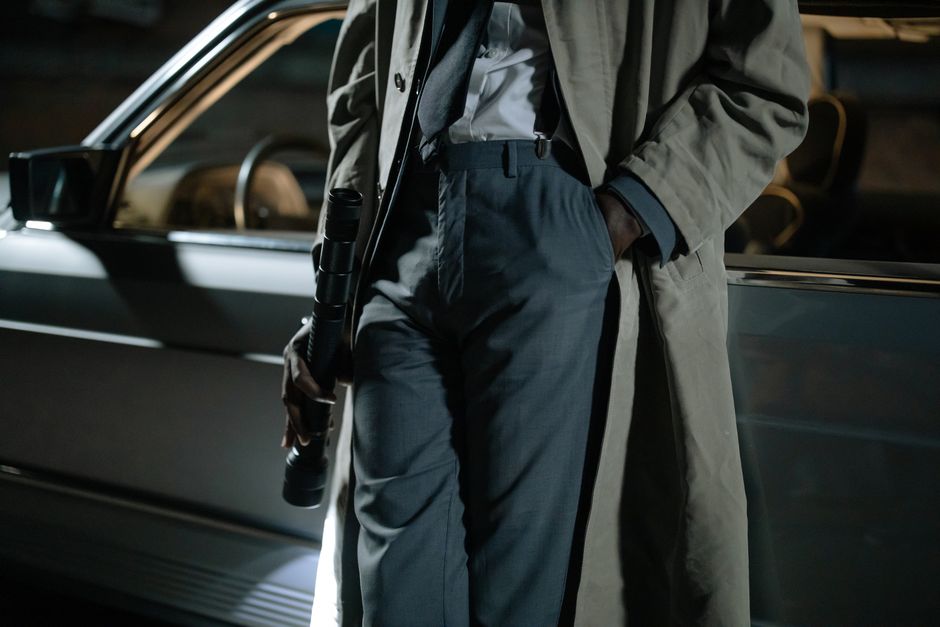 Close-up of a person in a trench coat holding a flashlight near a car at night.