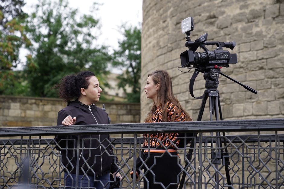 Two women conducting an outdoor interview near a historic stone wall, with a professional video camera set up on a tripod.