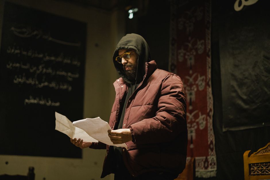 A man wearing a hooded jacket and glasses reads documents in a dimly lit room with decorative elements.