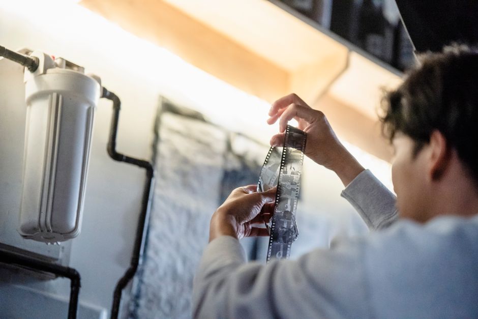 A person examines a strip of film in a darkroom setting with bright overhead lighting.