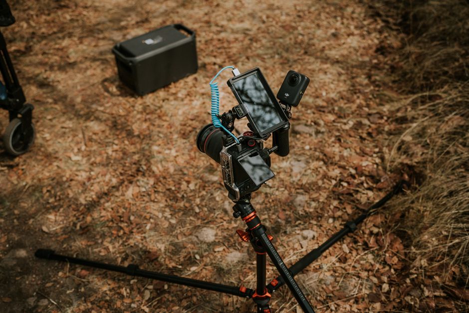 A camera tripod with video setup on a leaf-covered trail in an autumn forest setting.