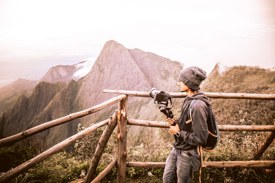 A young man with a camera stands at a wooden fence, photographing mountains.
