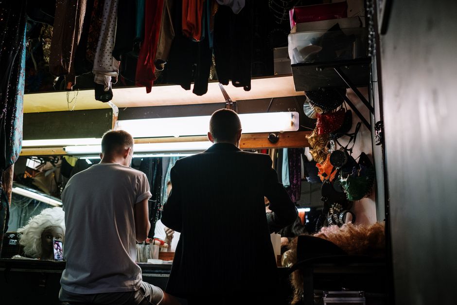 Two individuals preparing in a dimly lit dressing room with costumes and wigs, creating an artistic backstage ambiance.