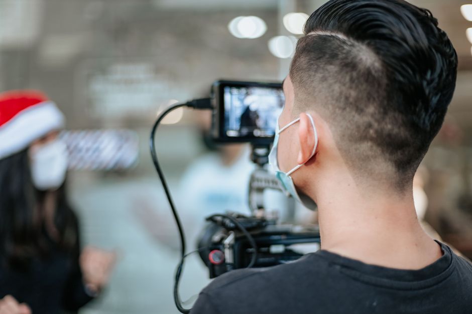 A masked videographer films an interview indoors with professional camera gear.