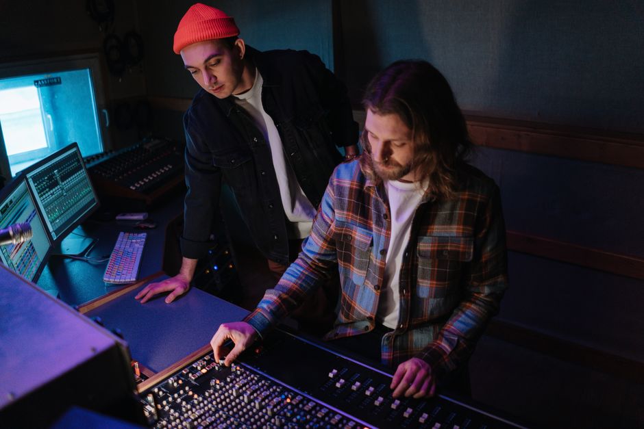 Two audio engineers mix sound on a console in a dimly lit, high-tech recording studio.