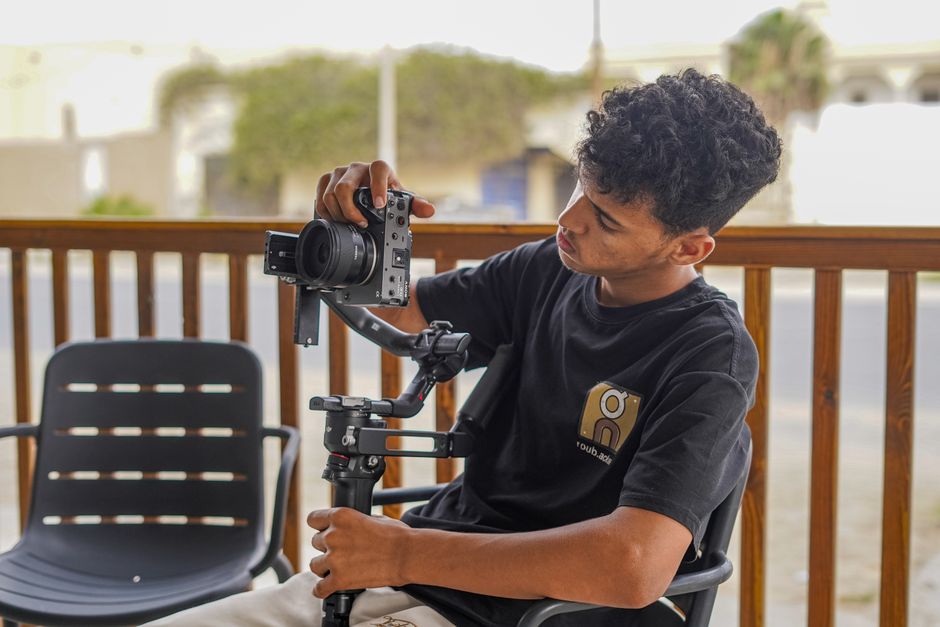 A young male photographer adjusts his camera gimbal on a patio chair outdoors.