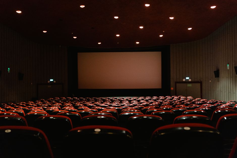 A dimly lit empty cinema theater with rows of red chairs facing a projector screen.