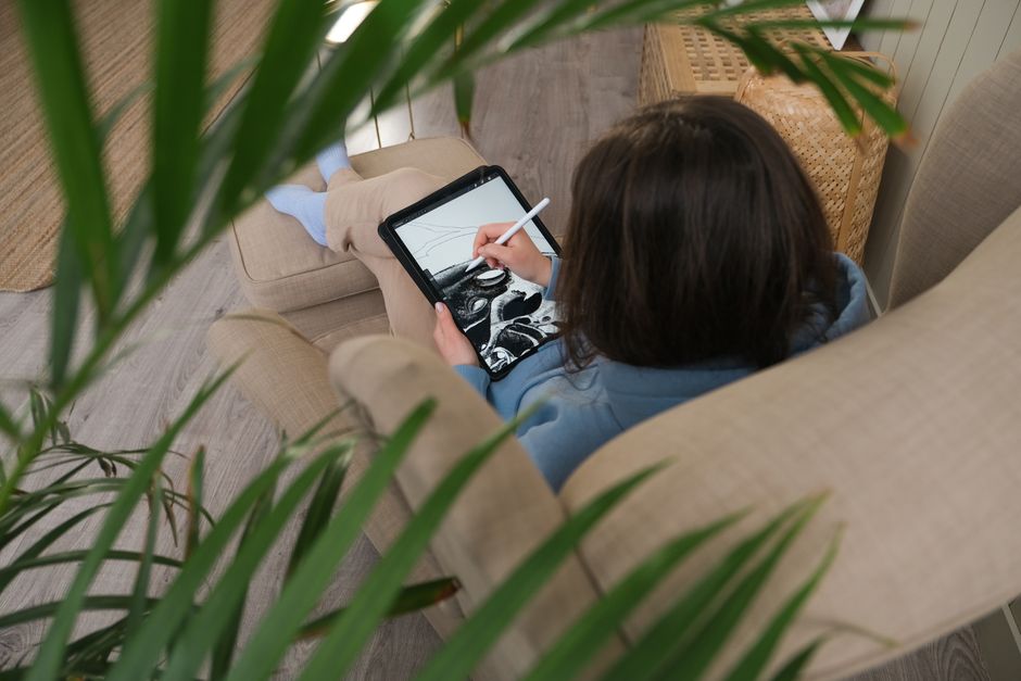A person drawing on a digital tablet while lounging on a sofa, surrounded by plant leaves.
