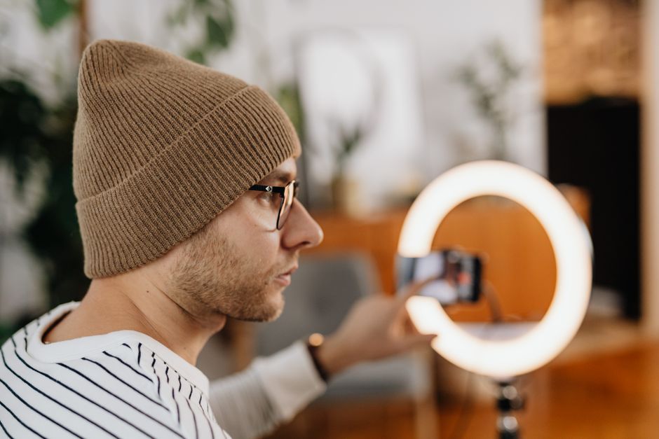 Man wearing a knitted cap uses a smartphone and ring light for video recording indoors.